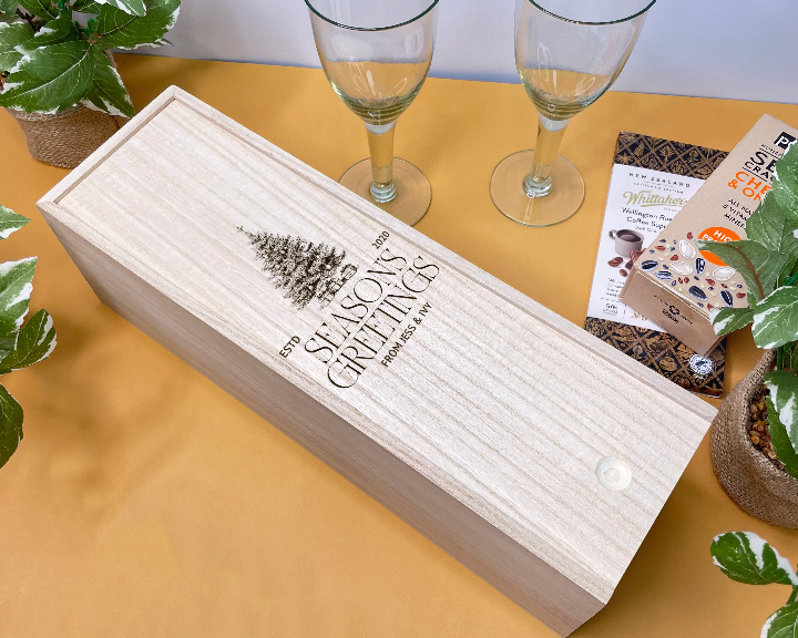 Wooden gift christmas wine box engraved with 'Season's Greetings' text on a table with wine glasses and plants.