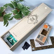 Wooden wedding wine box with tea bags, chocolate, and a seed cake on a white surface with plants in the background.